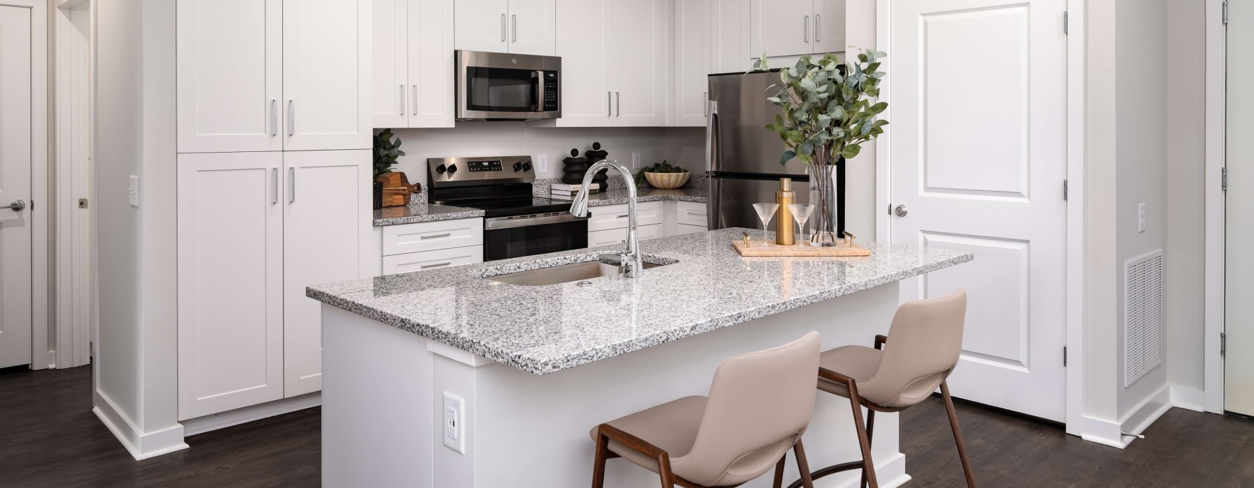 An interior view of a kitchen with a kitchen island countertop with a kitchen sink, two barstool chairs, kitchen cabinetry, and kitchen appliances, including a microwave, an oven, a fridge, and a stove in a one bedroom apartment for rent in Fairburn, GA a